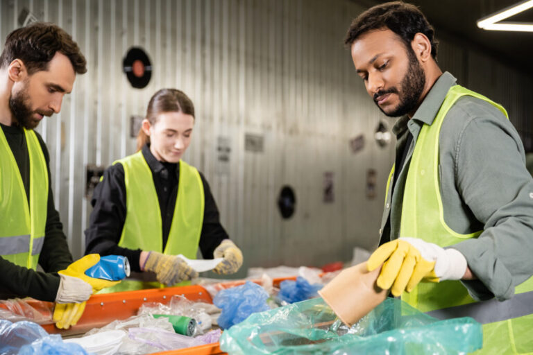 Bearded indian worker in safety vest and protective gloves putting paper cup in plastic bag while separating trash near conveyor and colleagues in waste disposal station, recycling concept